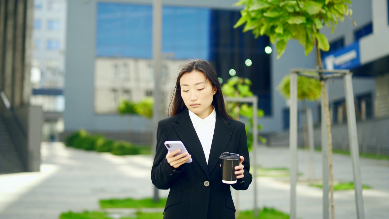 自信的经理商人走在城市街道附近的大现代商业区。年轻的亚洲女性用智能手机发短信，喝咖啡视频素材