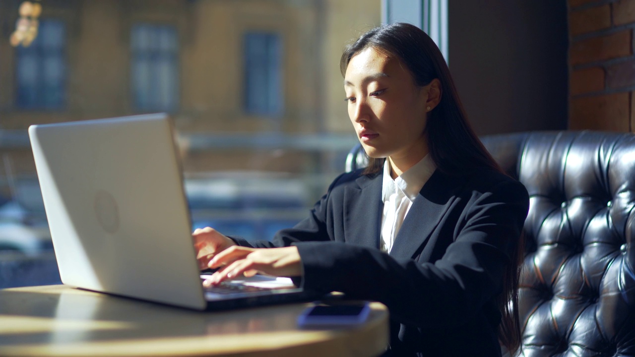 年轻的女性自由职业者或在线工作或在咖啡馆远程学习的学生。亚洲商业女性西装使用打字笔记本电脑在工作的共同工作热办公桌。视频素材