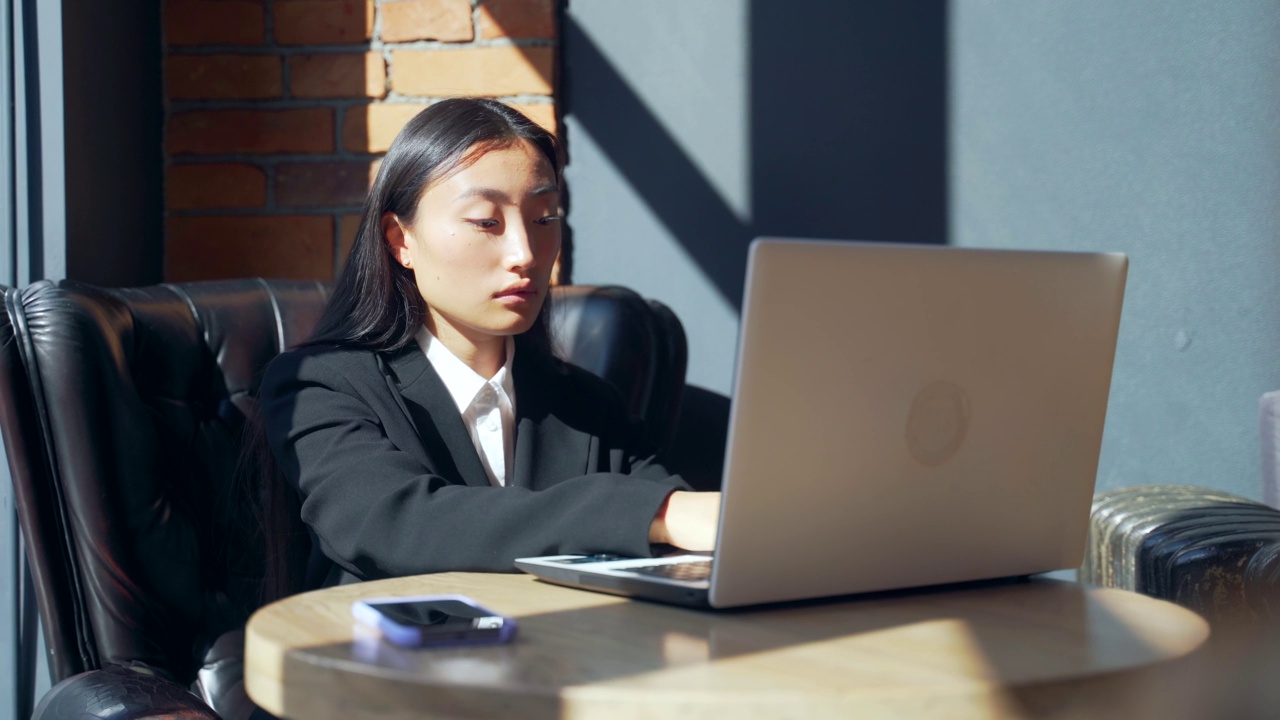 年轻的女性自由职业者或在线工作或在咖啡馆远程学习的学生。亚洲商业女性西装使用打字笔记本电脑在工作的共同工作热办公桌。视频素材