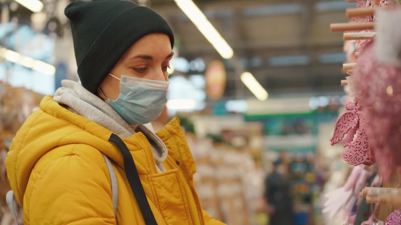 在购物中心的圣诞市场上选择圣诞饰品的黄色冬季夹克的女人。成年白种人女性在购买医用防护口罩过年装饰视频素材