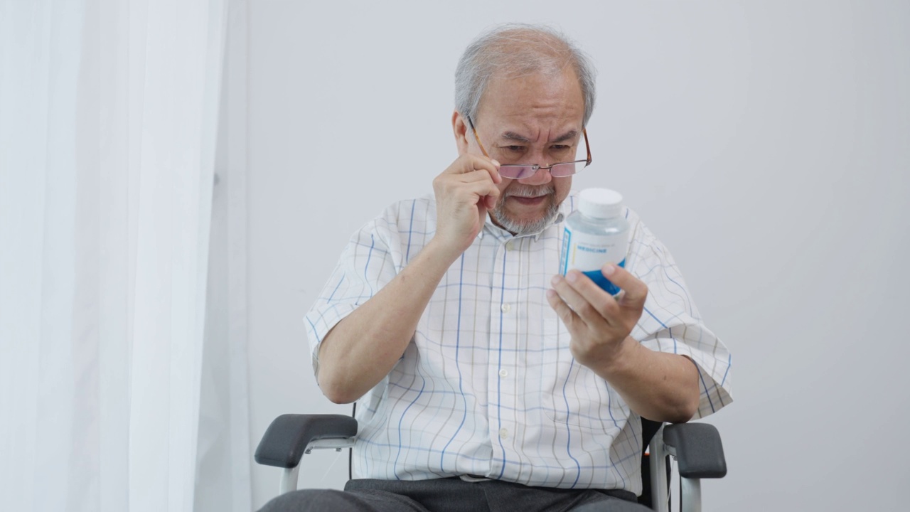 亚洲老年男性坐在轮椅上阅读处方。有魅力的老人，年长成熟的男子戴着眼镜，然后看药瓶在养老院。医疗保险的概念。视频素材