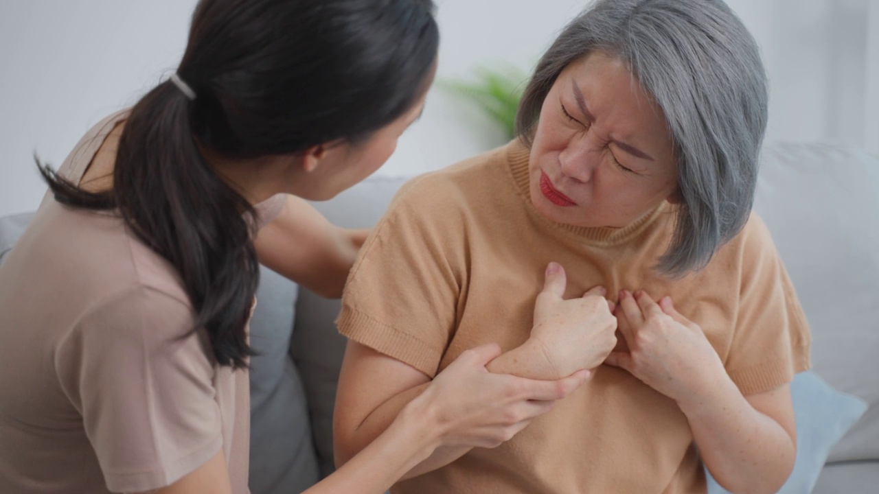 亚洲女儿帮助老年母亲心脏病发作胸痛。有魅力的女孩照顾和支持老年病人，她的胸部因剧烈疼痛而呼吸困难。视频素材