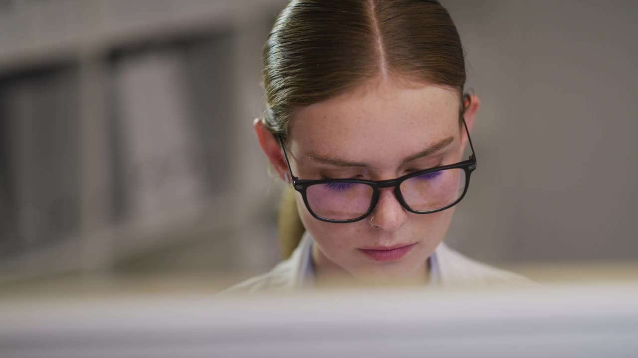 一位女科学家戴着眼镜坐在电脑前。年轻的职业女医生，在阅读和使用桌面显示器时戴眼镜矫正视力视频素材