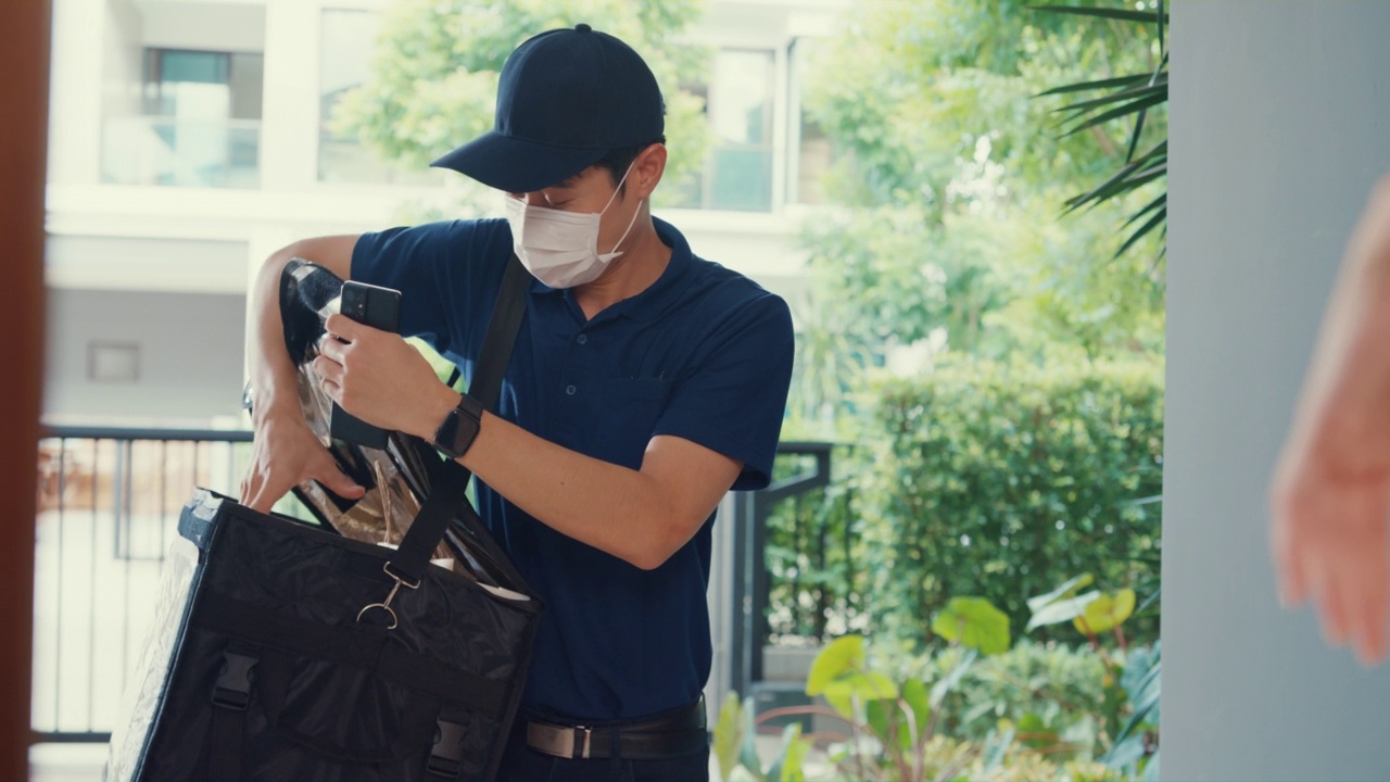 年轻的亚洲快递员戴着面具，穿着蓝色海军制服携带箱子敲门回家网上购物纸袋到女人在门前女士扫描二维码支付在家里的智能手机。视频素材