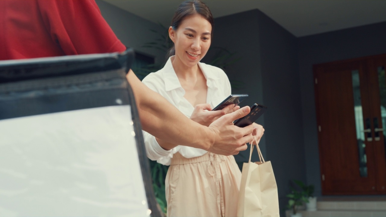 年轻快乐的亚洲女人接受纸袋从快递员与自行车在红色统一携带箱子，扫描二维码支付在房子前面的智能手机。视频素材