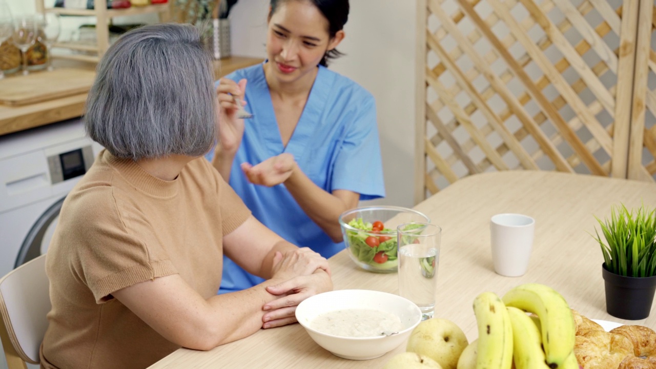 一位年轻的亚洲护士已经在家里准备好了饭菜，并在餐桌上给老祖母喂沙拉视频素材