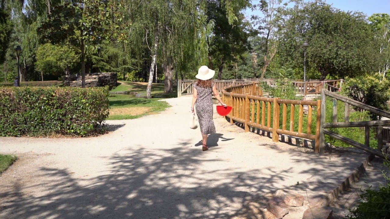 一个阳光明媚的夏日，一名女子穿着裙子，戴着草帽穿过公园。视频素材