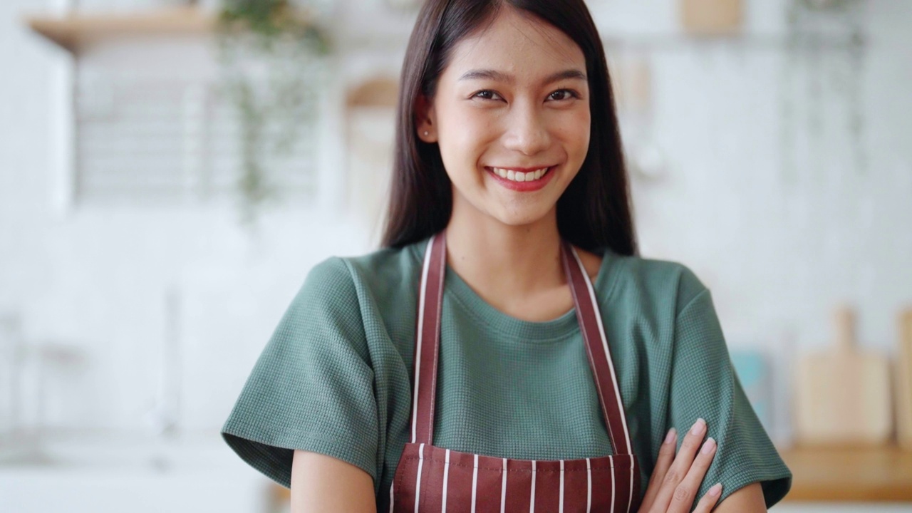 快乐的年轻亚洲女人穿着围裙，站在厨房的房间。美丽的女性微笑着看着镜头感觉自信视频素材