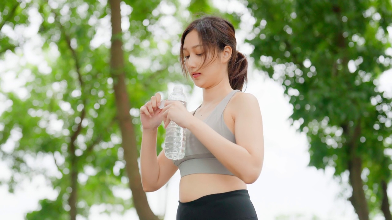 年轻的亚洲女性喝瓶装水，在她的锻炼期间休息。快乐的青春期女性在公园锻炼和休息休息喝水视频素材