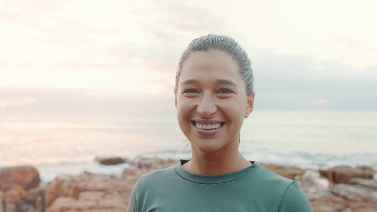 一个女人微笑着站在沙滩上的肖像。迷人的女性在海上看着镜头。快乐的女士在户外度假，享受她的假期。快乐的女士独自在岩石海岸视频素材