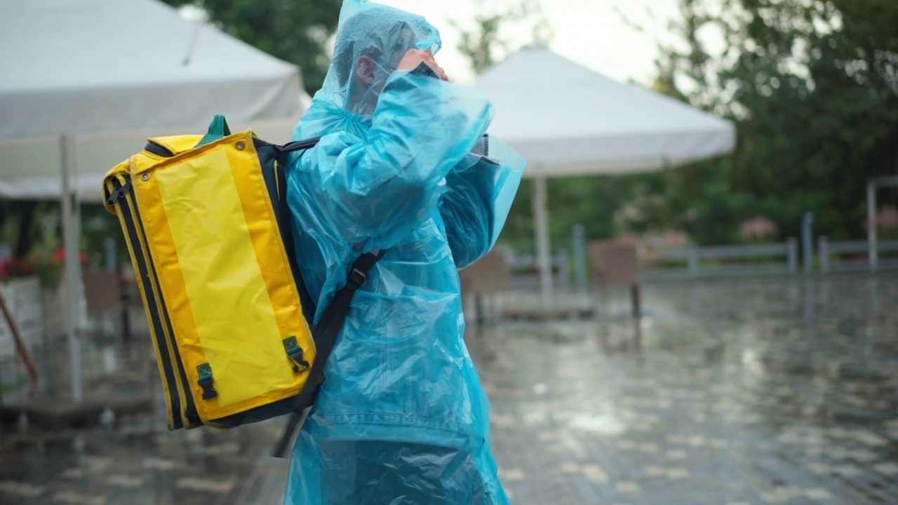 后视图快递员穿着雨衣和冠状病毒口罩，背着背包走在城市街道上四处张望。高加索快递员搜索地址在下雨的夏天送食物和饮料。视频素材