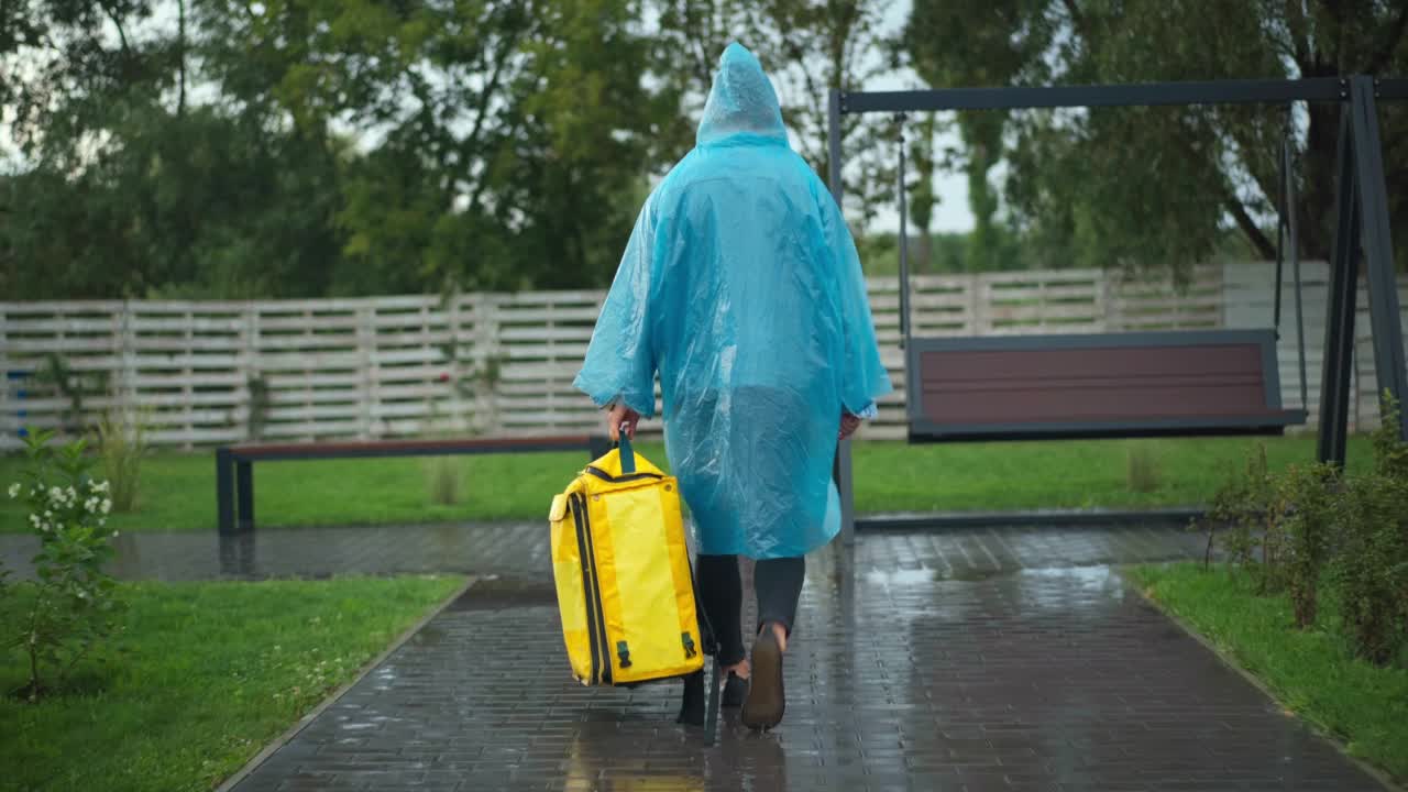 后视图广角镜头信使雨衣步行与黄色的背包在城市公园转向。直播镜头跟踪了一名在下雨天在户外送食物和饮料的年轻白人男子。视频素材