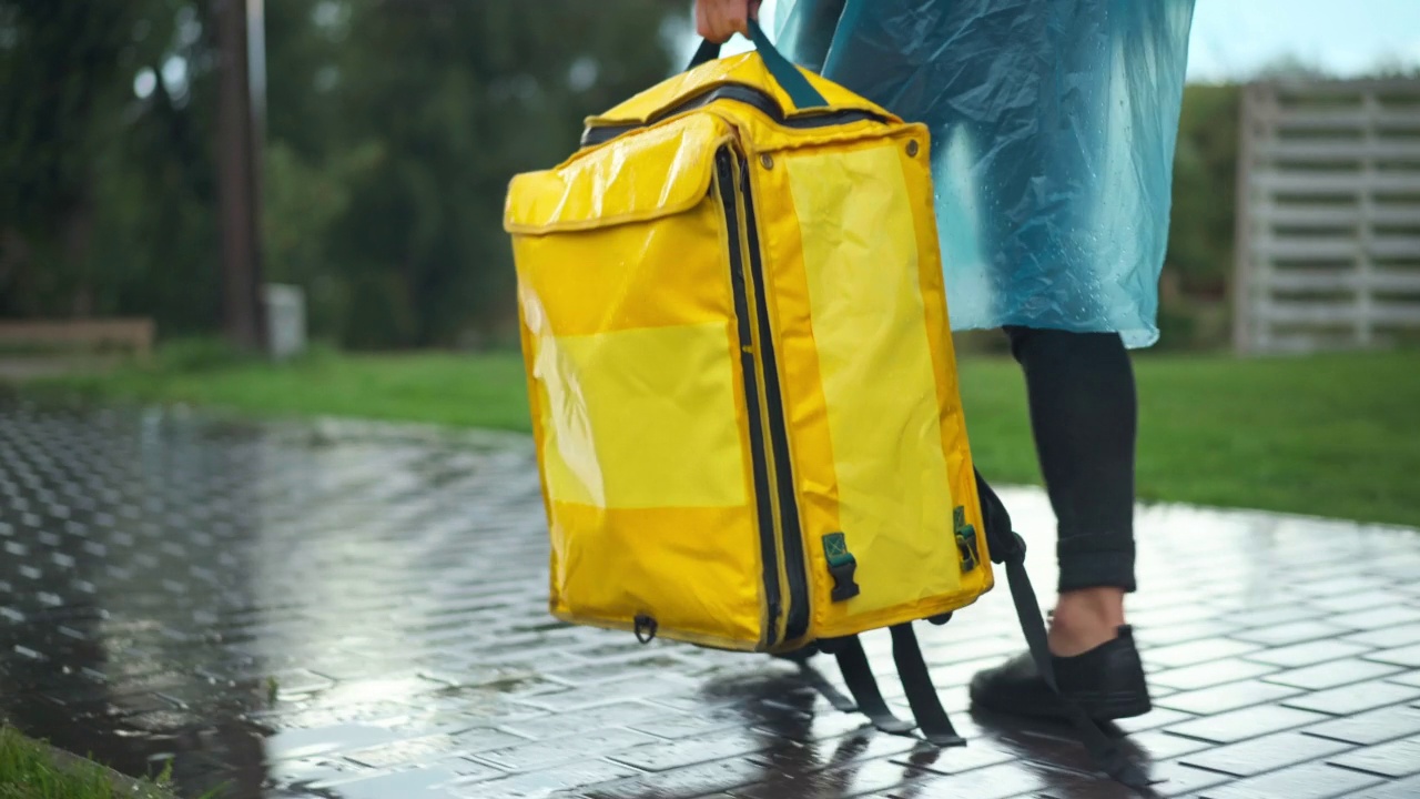 一名身着雨衣、面目全非的快递员手拿黄色快递包在城市公园散步的跟踪镜头。年轻的白人男子背着背包在户外的人行道小巷里散步。服务和天气。视频素材