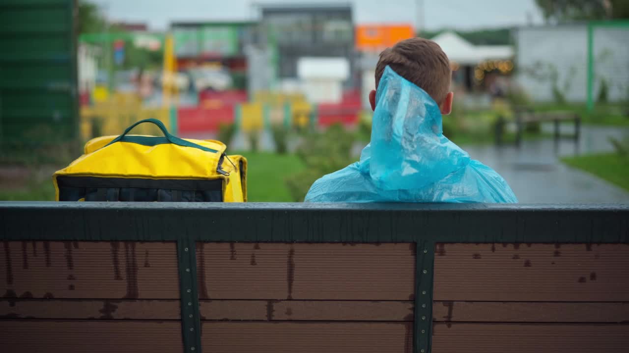 背黄色背包的年轻快递员坐在城市街道的长凳上。雨后穿着雨衣的白人男快递员在城外等客户。视频素材