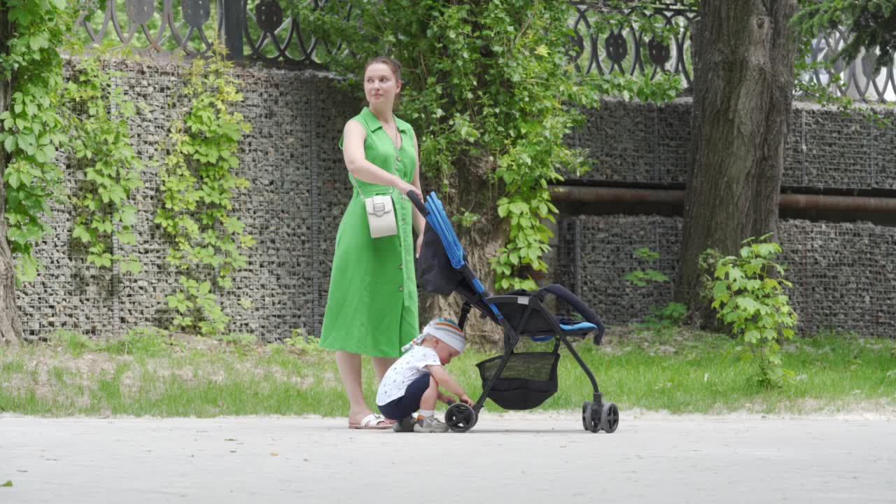 夏天，妈妈带着孩子在公园里散步，阳光明媚的日子里，孩子推着婴儿车在人行道上和妈妈玩耍。视频素材