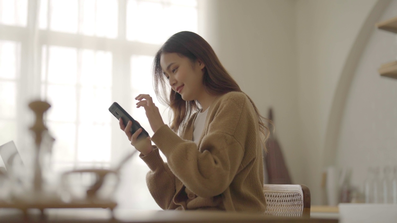 快乐的年轻亚洲女人在家里放松。亚洲女性站在厨房柜台，使用手机视频素材