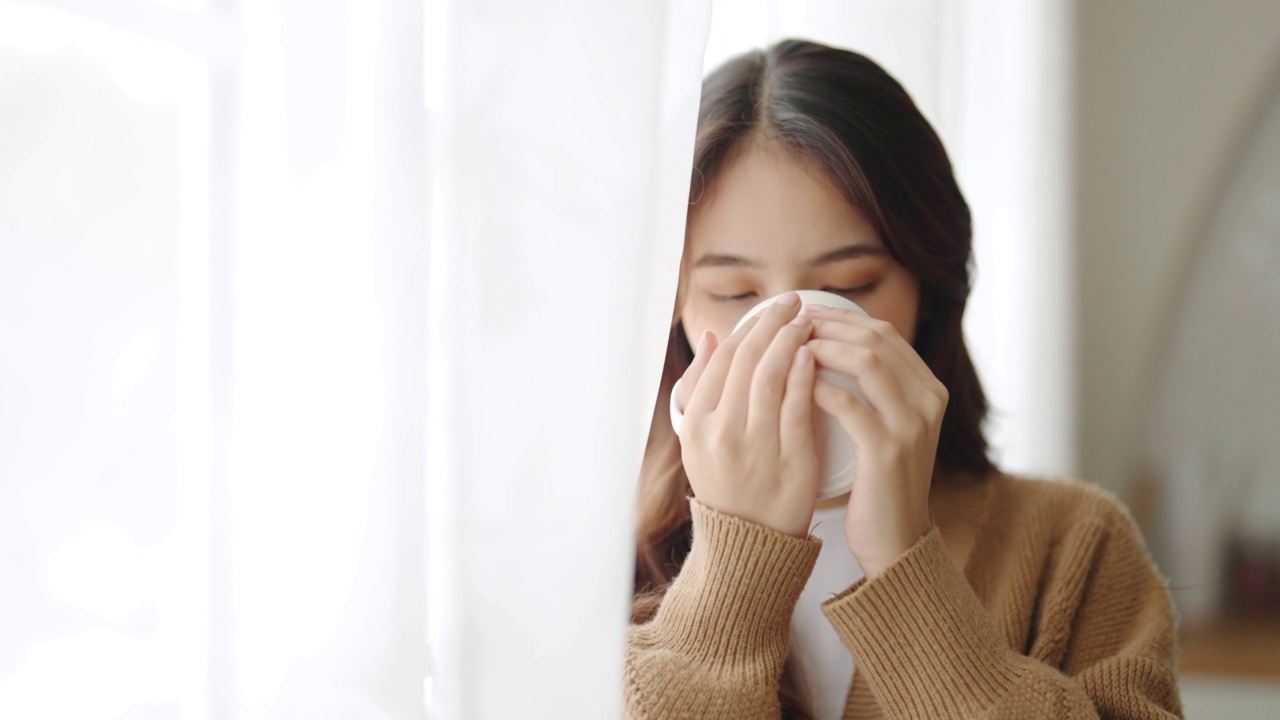 年轻的亚洲女性站在窗边，拿着杯子在家里的客厅里。亚洲女性早上醒来后喝牛奶或茶视频素材