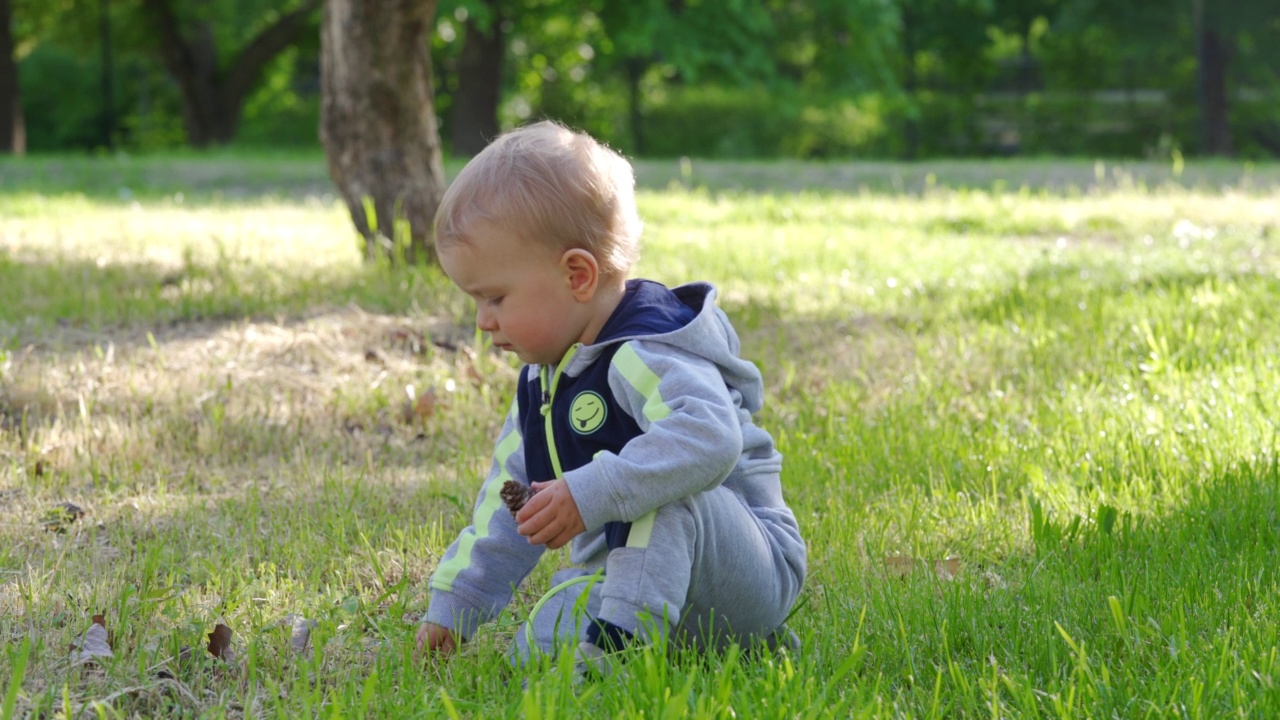 孩子坐在春天公园的绿草地上，1.5岁的幼童在绿草地上玩耍。儿童健康的夏季活动。视频素材