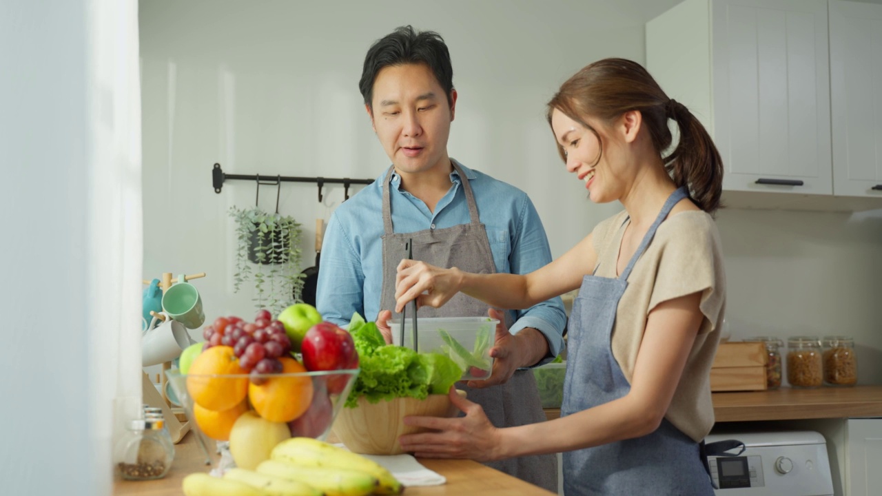 幸福的家庭，亚洲迷人的夫妇在家里的厨房做饭。年轻男女穿围裙，花时间做健康沙拉早餐，在家保健。饮食与健康食品理念视频素材