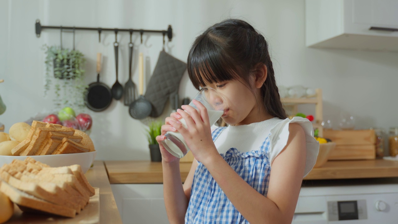 亚洲小孩拿着一杯牛奶在家里的厨房里喝。幼小的学龄前女孩或女儿喝牛奶，弄乱了牛奶的胡子和嘴唇后，在家里的医疗保健。视频素材