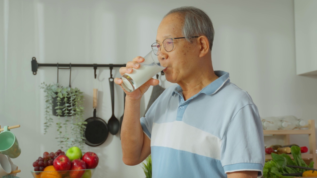 亚洲高级成熟男性在家里的厨房喝一杯牛奶。有魅力的老爷爷抱着一杯牛奶，喝着，早上起床后在家里做保健。视频素材