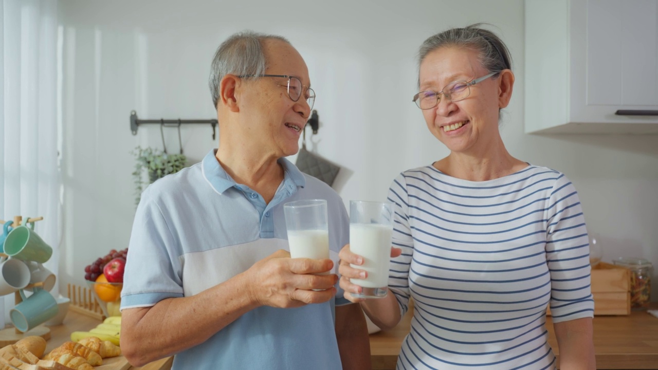 亚洲老夫妇在家里的厨房里喝一杯牛奶的肖像。有吸引力的年长的年长的祖父母抱着和啜饮一杯牛奶后，早上醒来在家里的保健视频素材