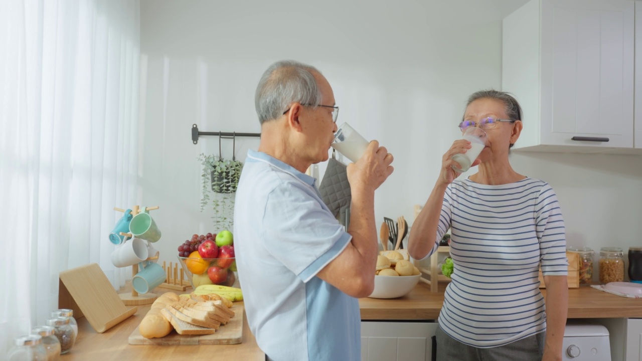 亚洲资深成熟夫妇在家里的厨房喝一杯牛奶。有吸引力的年长的年长的祖父母抱着和啜饮一杯牛奶后，早上醒来在家里的保健。视频素材