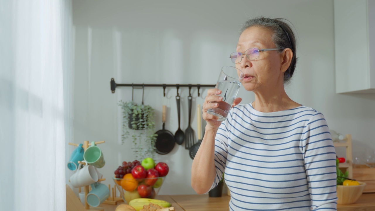 亚洲高级成熟女性在家里的厨房喝一杯牛奶。有魅力的老祖母抱着和喝着一杯牛奶，早上醒来在家里的保健。视频素材