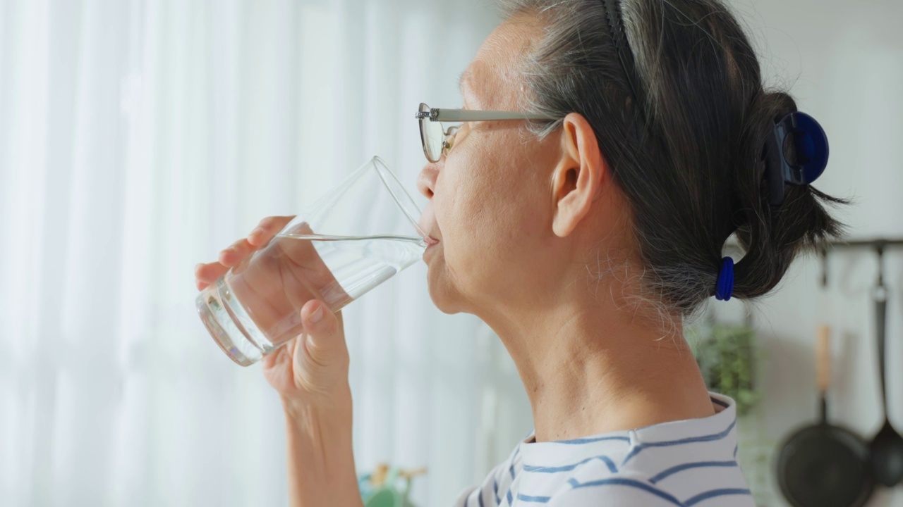 亚洲高级成熟女性在家里的厨房喝一杯水。有吸引力的老祖母抱着清洁的矿物自然在杯子后，早上醒来在家里的健康福祉视频素材