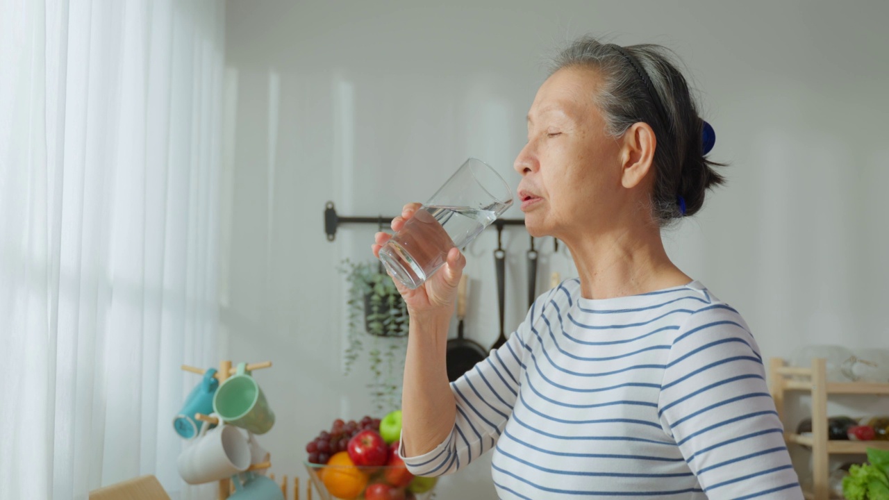 亚洲高级成熟女性在家里的厨房喝一杯水。有吸引力的老祖母抱着清洁的矿物自然在杯子后，早上醒来在家里的健康福祉视频素材