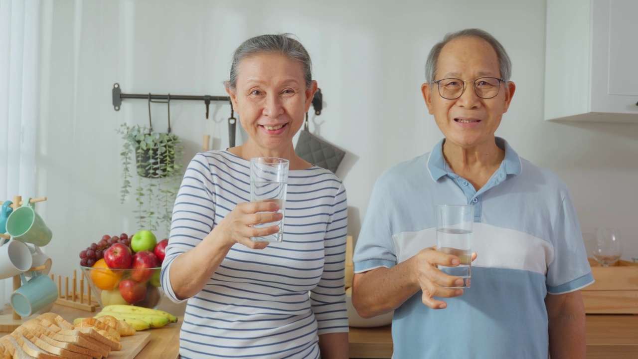 亚洲老夫妇在家里的厨房里喝一杯水的肖像。有吸引力的年长的祖父母抱着清洁的天然矿物在杯子后，早上醒来在家里的健康福祉视频素材