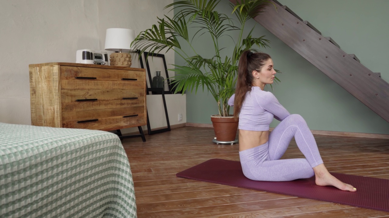 一位穿着紧身紫色西装的女士在家里做瑜伽。坐在地毯上，做一个体式。双腿扭曲，身体向一侧倾斜。热身，拉伸，体操，运动，健康的生活方式视频素材