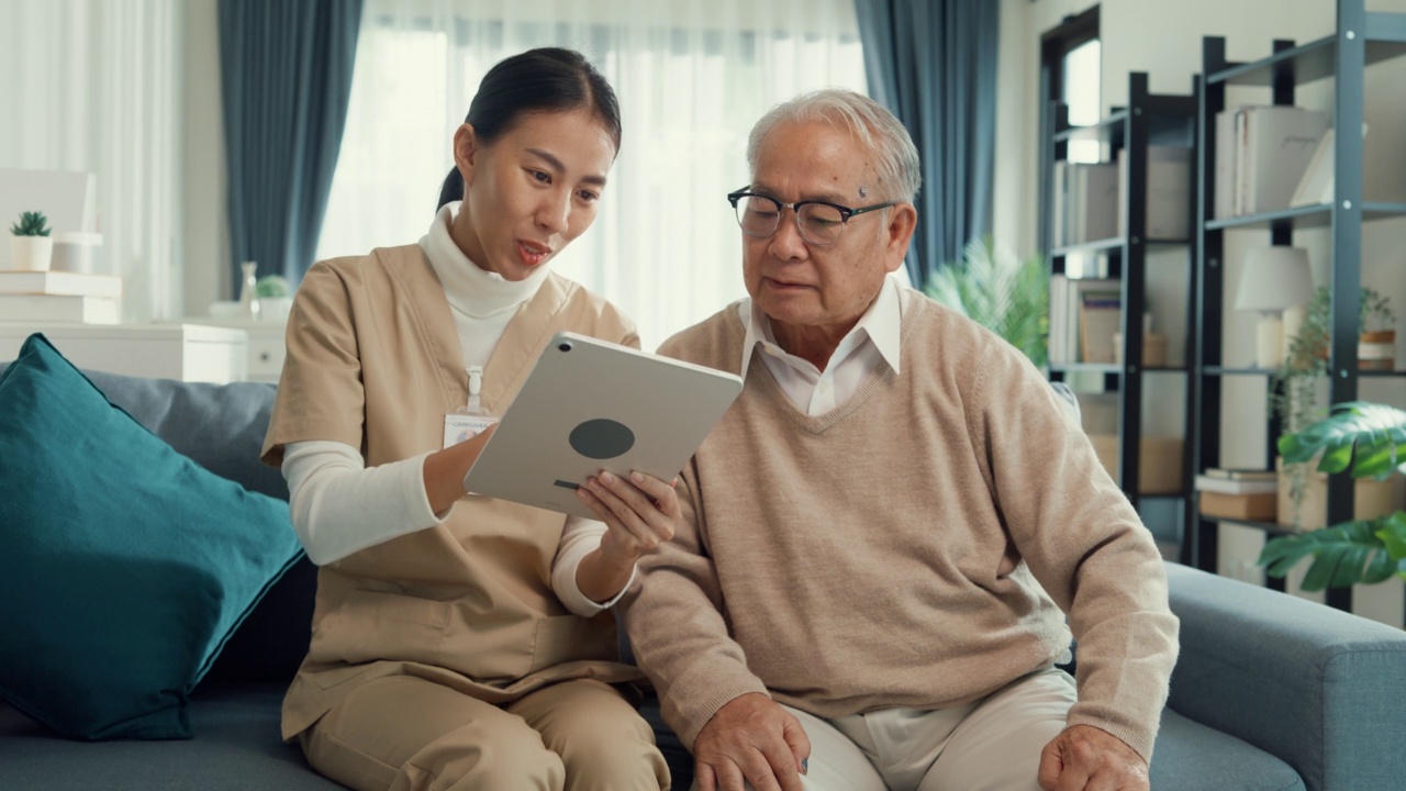 年轻的亚洲女性专业护理人员，个人护理护士教年长的男人在家里的沙发上使用数字平板电脑。视频素材