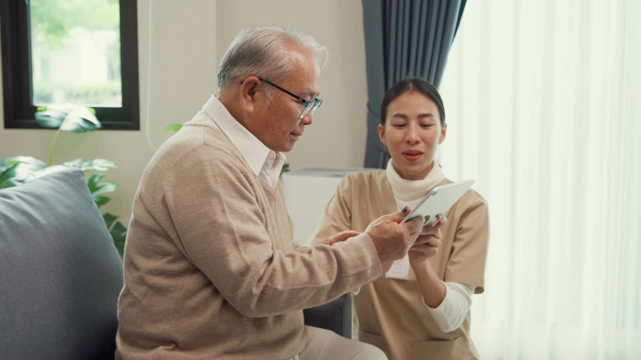 年轻的亚洲女性专业护理人员，个人护理护士教年长的男人在家里的沙发上使用数字平板电脑。视频素材