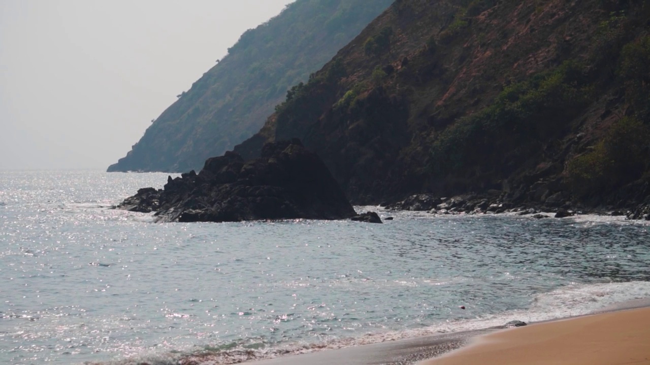 从印度果阿的Kakolem海滩看到的岩石山前面的海浪的慢动作拍摄。热带海滩岛屿背景。山边的沙滩上海浪滚滚。自然背景。视频素材