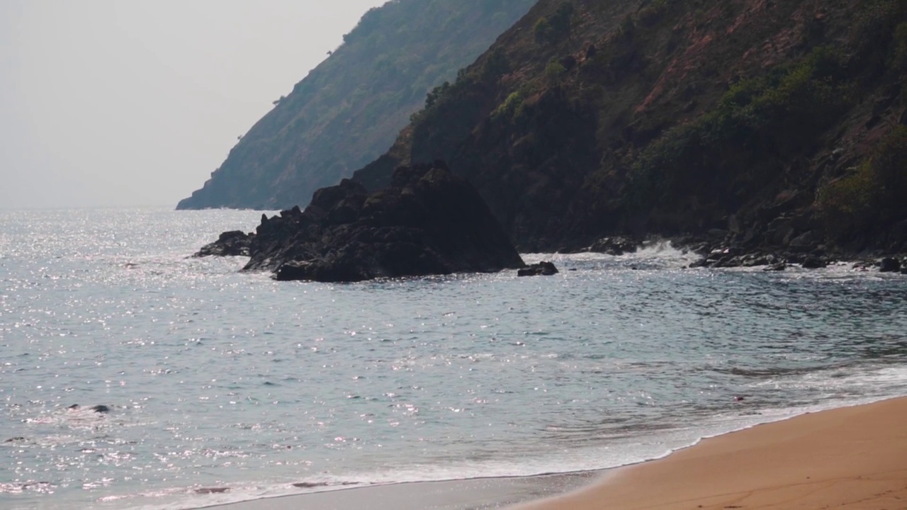 从印度果阿的Kakolem海滩看到的岩石山前面的海浪的慢动作拍摄。热带海滩岛屿背景。山边的沙滩上海浪滚滚。自然背景。视频素材