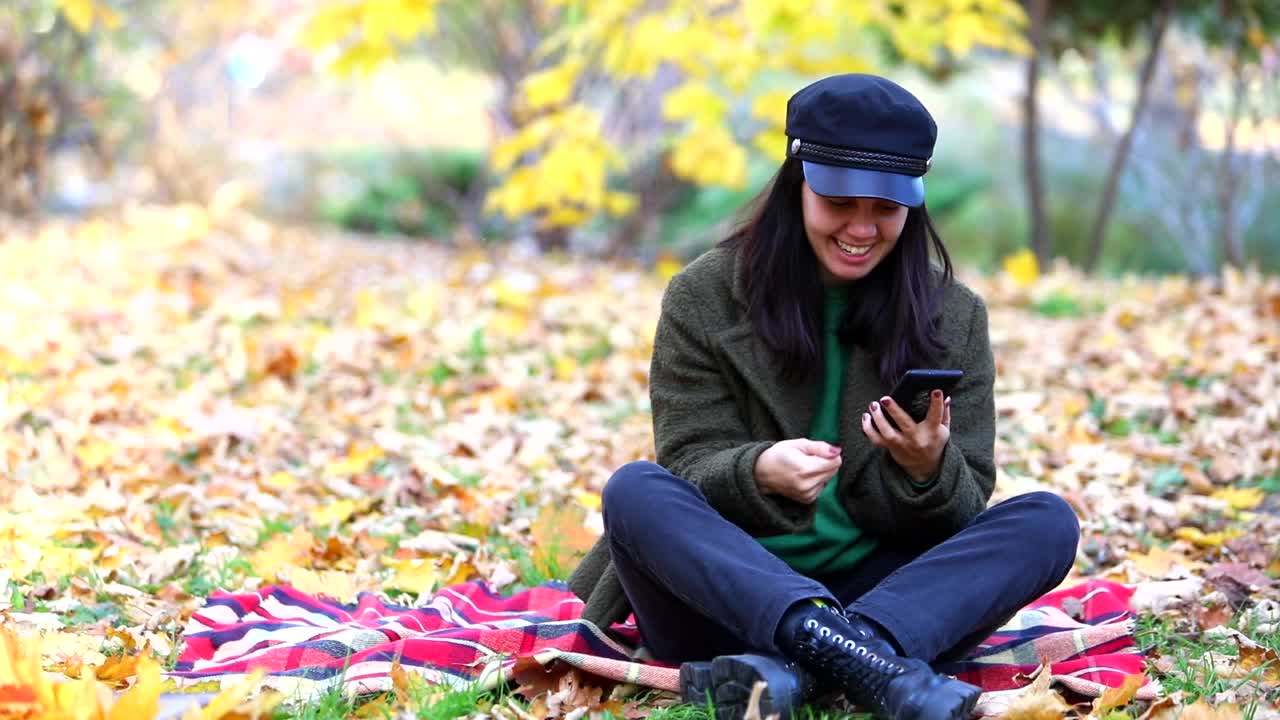 快乐女人拿着手机坐在秋天公园的毯子上视频素材