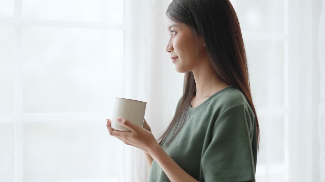 年轻的亚洲女子站在窗边，拿着杯子在家里的卧室，她早上醒来后喝牛奶视频素材