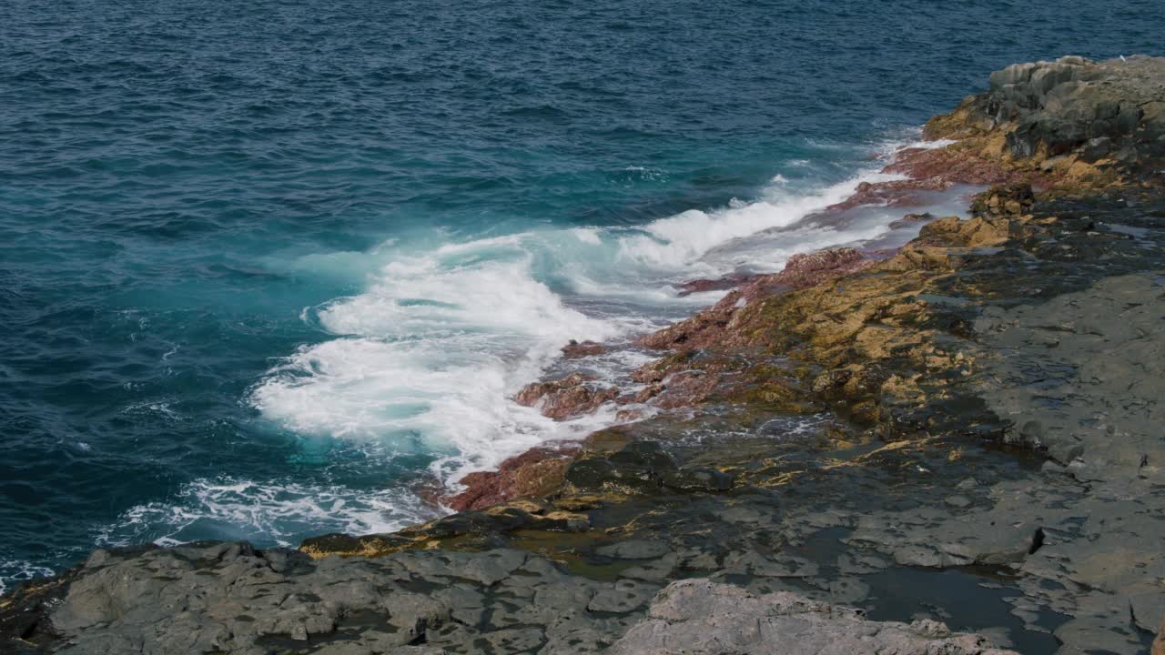 海浪冲刷着黑色的火山岩岩石。岸上溅起的浪花，水面的反光。阳光明媚的一天。视频素材