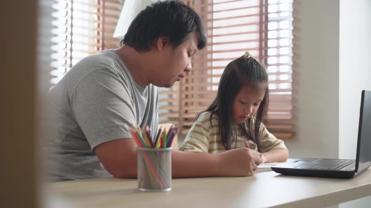 亚洲单身父亲在家里的客厅用笔记本电脑学习在线电子学习，并在书上写字，以此来教育和照顾女儿。快乐快乐的亲子关系在居家教育活动中的新常态概念。视频素材