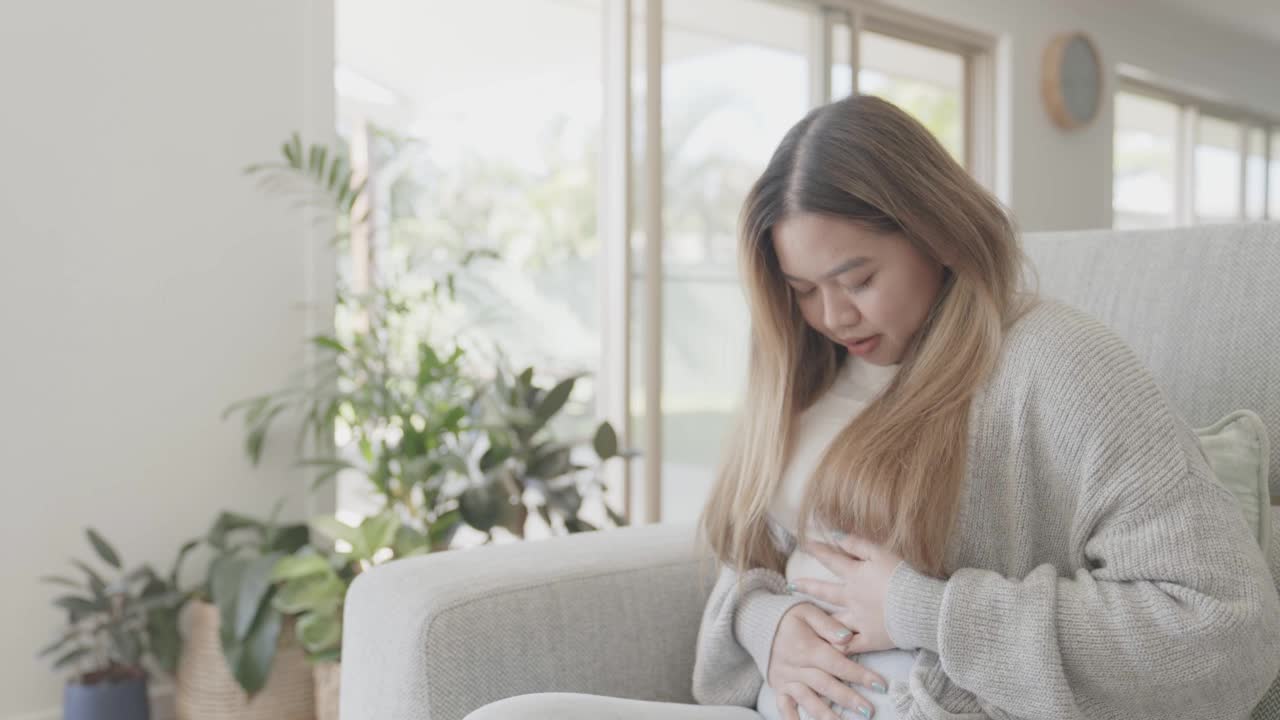 年轻的亚洲女性躺在家里的沙发上，肚子疼，有肠道保健的概念视频下载