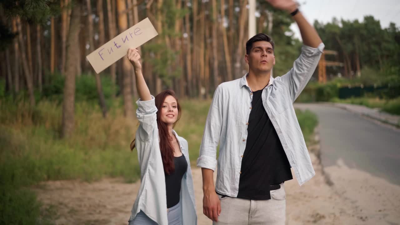 快乐的年轻人和女人搭便车与未来的旗帜微笑交谈。积极的白人男女朋友站在郊区公路上的森林边。旅行和旅游。视频素材