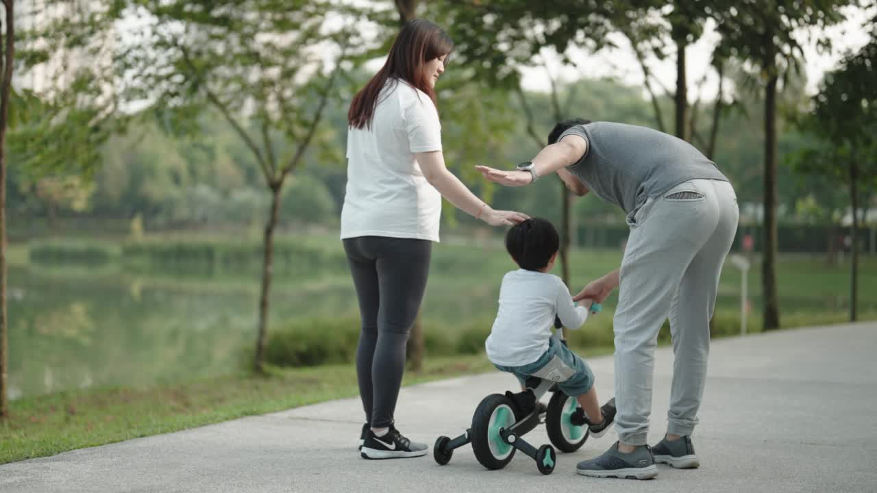 亚洲华裔男孩在周末早上和他的父母在公园里学习骑自行车视频素材