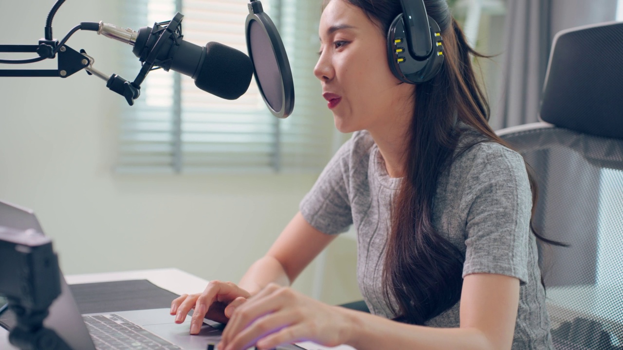 亚洲迷人的音频DJ女人对着麦克风讲话广播。年轻漂亮的女博主网红戴着耳机，在家录音棚为电台录制早间新闻播客节目。视频素材