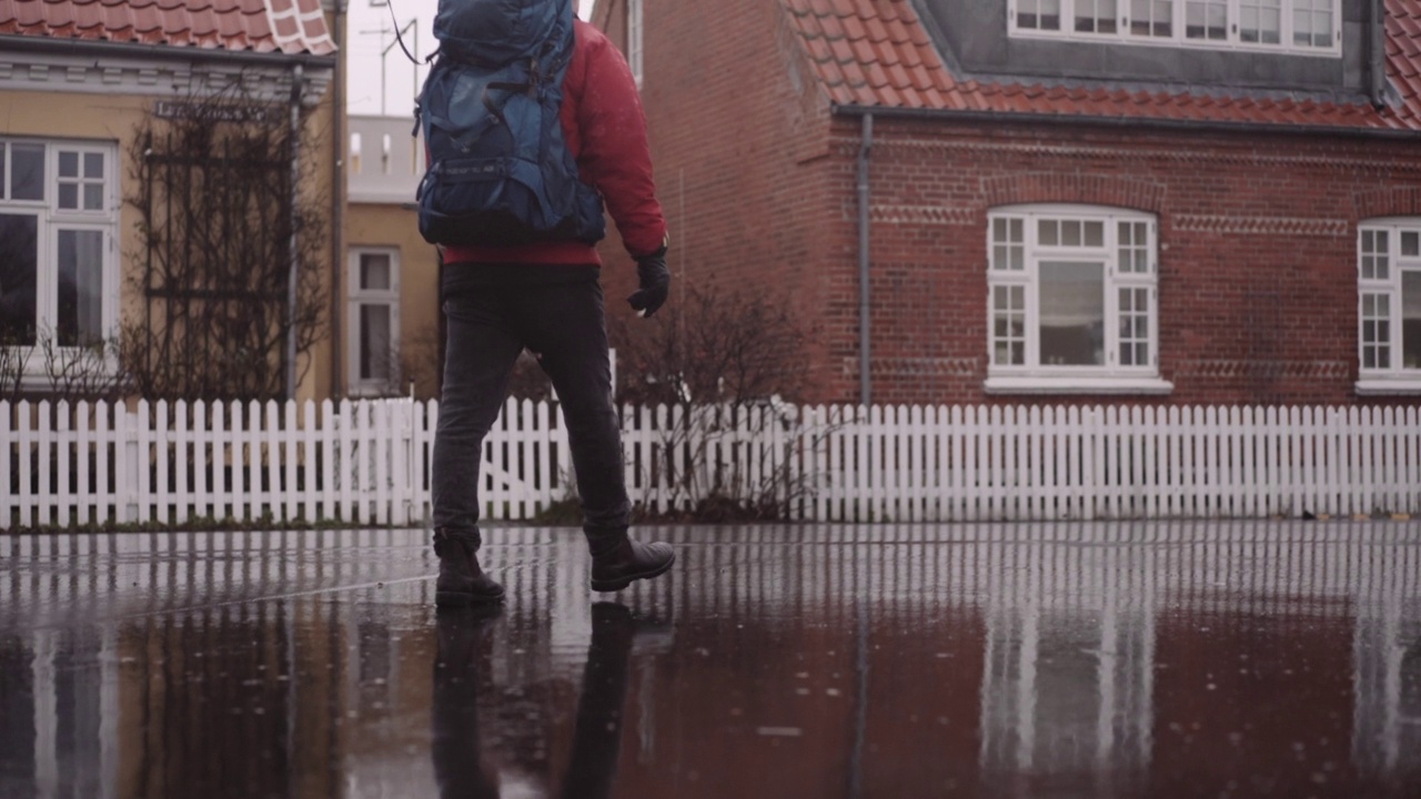 穿着冬装的背包男子在雨中走在潮湿的柏油路上视频素材