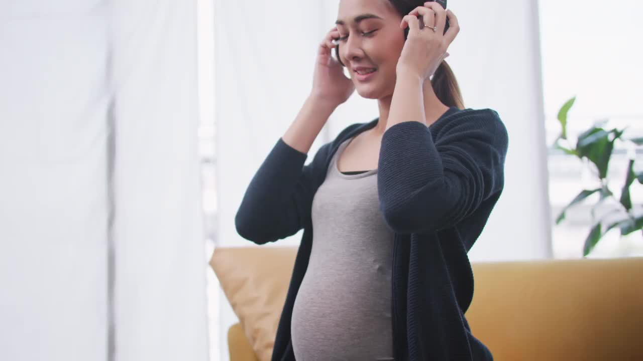 快乐的亚洲孕妇把耳机戴在肚子上。视频素材