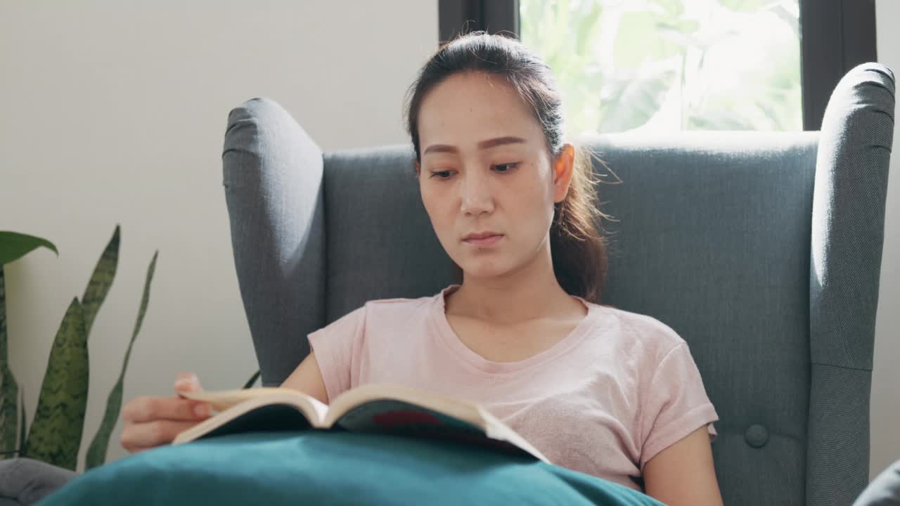 年轻的亚洲大学女子与休闲坐在翼椅上阅读最喜欢的书文本学习在家里的客厅。视频素材