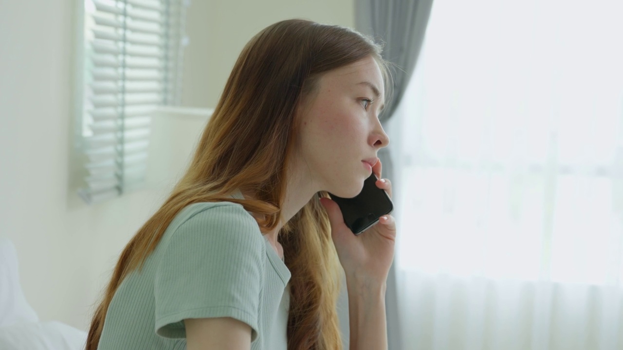 沮丧的白人妇女在家里的卧室里打电话。迷人的年轻女孩把闲暇时间花在床上，在家里用智能手机交流和讨论时感到生气和不高兴。视频素材