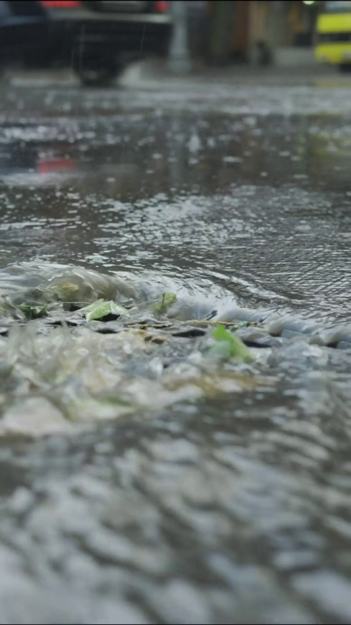 垂直视频:特写雨水流入雨水排水沟的场景，背景中是正在行驶的汽车。城里下起了暴雨。慢动作视频素材