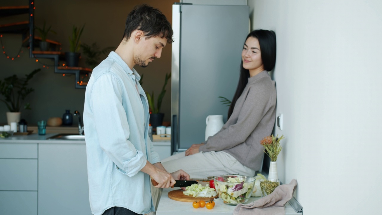 男人在做饭切蔬菜做沙拉，而女人坐在厨房的桌子上视频素材